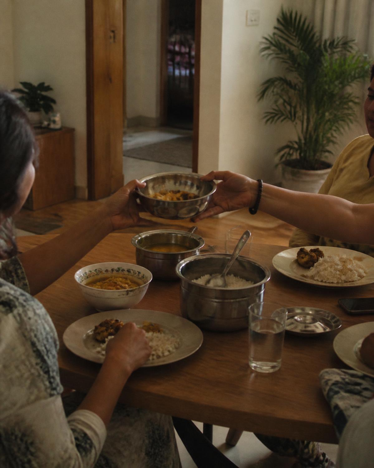 Guests passing a steel bowl of food across a small dining table in an Indian home